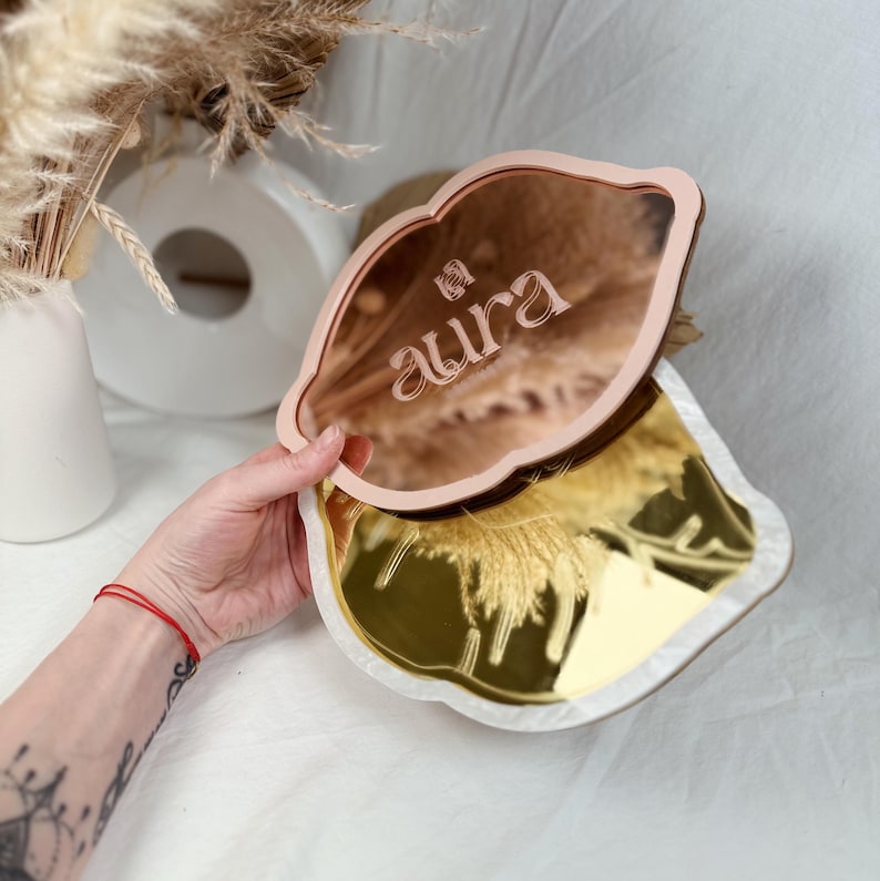 May include: Two decorative mirrors in the shape of a lip. The top mirror is pink with the word "aura" engraved on it. The bottom mirror is gold.