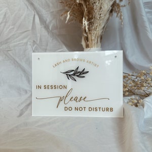 May include: White acrylic sign with black text that reads "Lash and Brow Artist" with a small black branch design. The sign also reads "In Session Please Do Not Disturb" in gold text.