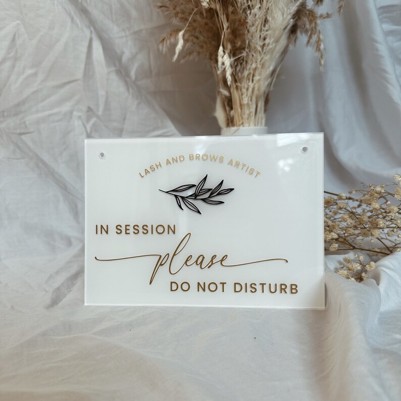 May include: White acrylic sign with black text that reads "Lash and Brow Artist" with a small black branch design. The sign also reads "In Session Please Do Not Disturb" in gold text.
