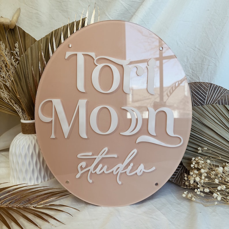 May include: A round, light pink acrylic sign with the text "Tori Moon Studio" in white lettering.