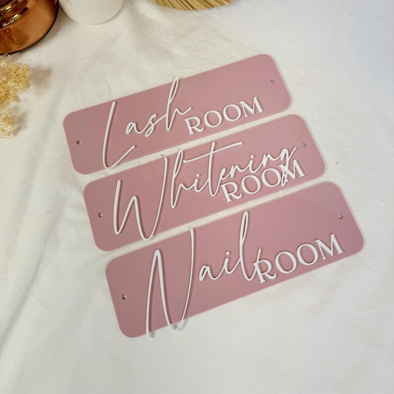 Op de afbeelding: Drie roze acrylplaten met witte letters. De platen lezen "Lash Room", "Whitening Room" en "Nail Room".