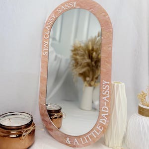 May include: Oval-shaped pink mirror with the text "STAY CLASSY, SASSY & A LITTLE BAD-ASSY" in white. The mirror is surrounded by a pink marbled frame. A candle and vases are in the background.