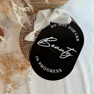 May include: A black oval sign with the words "DO NOT DISTURB Beauty IN PROGRESS" in white script. The sign is attached to a wooden round with a white ribbon.