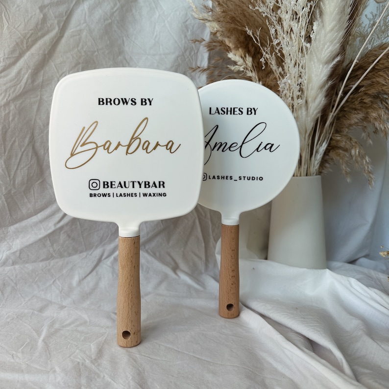Peut inclure: Deux miroirs &agrave; main blancs avec des manches en bois. L'un est carr&eacute; avec l'inscription "Brows by Barbara" et "BEAUTYBAR". L'autre est rond avec l'inscription "Lashes by Amelia" et "LASHES_STUDIO".
