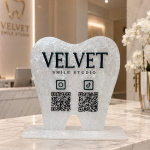 May include: A white, pearlescent, tooth-shaped acrylic sign featuring the black text VELVET SMILE STUDIO, an Instagram icon, a TikTok icon, and two distinct QR codes. The sign stands on a rectangular base and displays branding information.