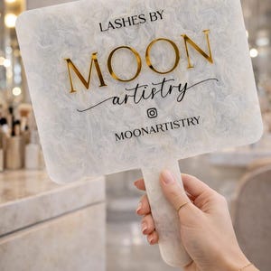 May include: A white hand mirror with a marbled design and a handle. The mirror features the text "LASHES BY MOON artistry" in gold lettering. The Instagram handle "MOONARTISTRY" is also displayed.