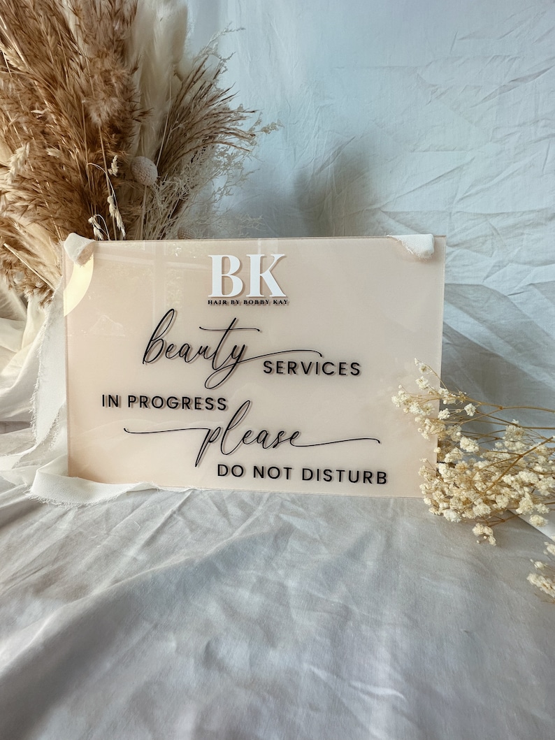 May include: A pink acrylic sign with black text that reads "BK Hair by Bobby Kay beauty services in progress please do not disturb".