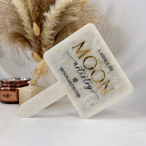 May include: A white and gold Moon Artistry lash brush with a handle. The brush has the text "Lashes by Moon Artistry" in gold lettering. The brush is on a white surface with a blurred background of dried flowers.