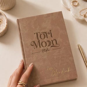 May include: A tan-colored journal titled "Tori Moon Studios" and "The Glow Book" with gold lettering. The journal is held by a person. Gold pens and a ring are visible on the table.