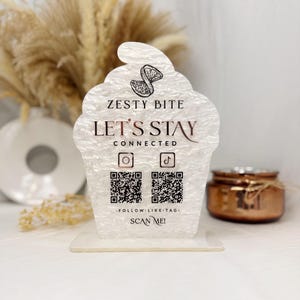 May include: A clear, cupcake-shaped sign with the text "Zesty Bite" and "Let's Stay Connected." It features a lemon slice graphic and social media icons with QR codes. The sign is on a clear base.