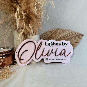 May include: Pink and white sign with the text "Lashes by Olivia" in gold lettering. The sign also includes the Instagram handle "@OliviaBeauty".