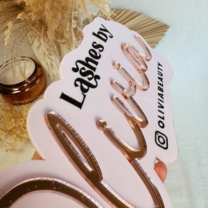 May include: Pink acrylic sign with the text "Lashes by" in black and "Olivia" in rose gold lettering. The sign has a wavy edge and the Instagram handle "@OliviaBeauty" is printed in black in the bottom right corner.
