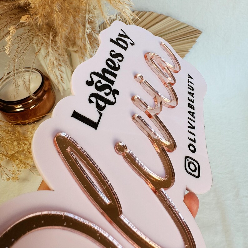 May include: Pink acrylic sign with the text "Lashes by" in black and "Olivia" in rose gold lettering. The sign has a wavy edge and the Instagram handle "@OliviaBeauty" is printed in black in the bottom right corner.