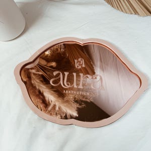 May include: A pink, lip-shaped, decorative mirror with a rose gold backing. The mirror has the text "aura aesthetics" engraved on the surface.