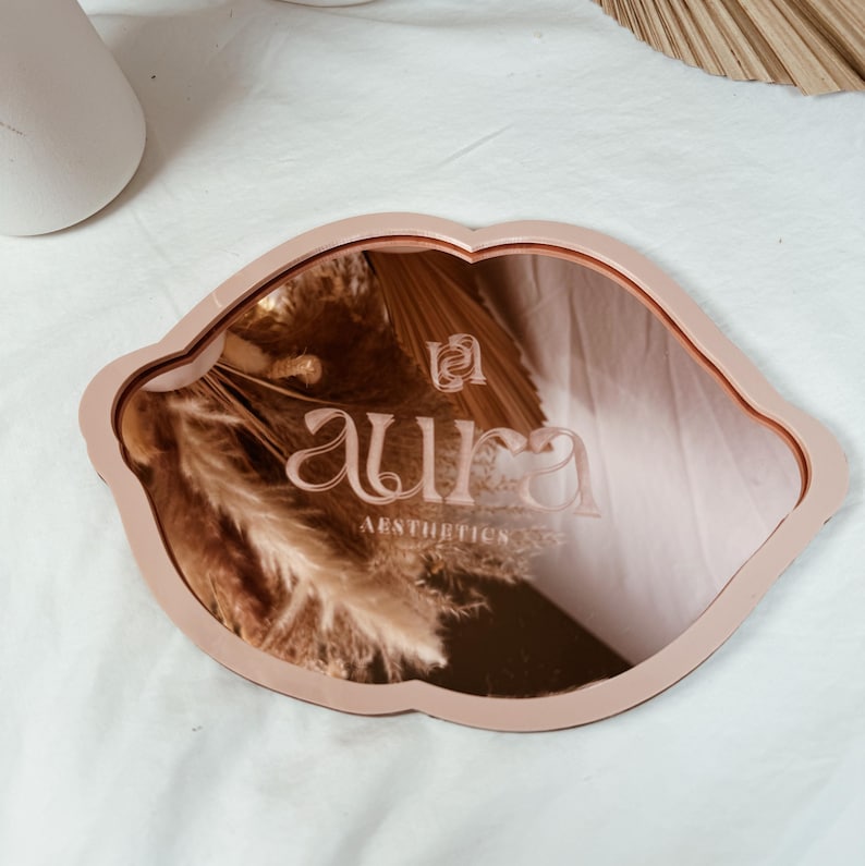 May include: A pink, lip-shaped, decorative mirror with a rose gold backing. The mirror has the text "aura aesthetics" engraved on the surface.