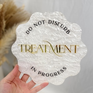 May include: A white, scalloped-edge sign with the text "DO NOT DISTURB TREATMENT IN PROGRESS" in black and gold lettering. The sign has a textured, marbled appearance and a small hole at the top for hanging.