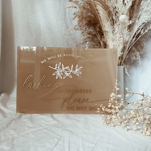 May include: A brown acrylic sign with gold lettering that reads "We will be right with you. Lashing in progress please do not disturb". The sign has a white floral design.
