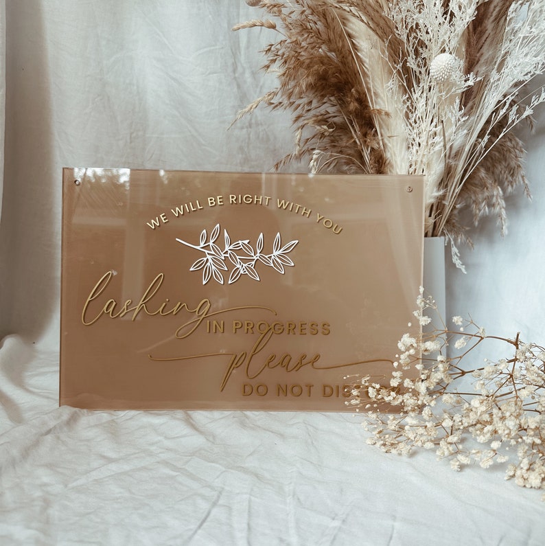 May include: A brown acrylic sign with gold lettering that reads "We will be right with you. Lashing in progress please do not disturb". The sign has a white floral design.