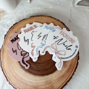 May include: Three decorative nail art business coasters with scalloped edges, each featuring the words "NAILS" and "Nails" in different colors. The coasters are arranged on a wooden slice.
