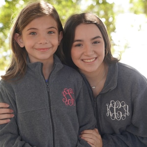 Puede incluir: Dos chicas con chaquetas de forro polar grises con monogramas bordados. La chica de la izquierda tiene un monograma rosa y la chica de la derecha tiene un monograma blanco.