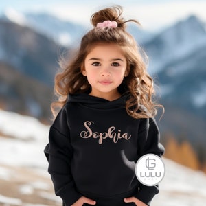 May include: A black hoodie with a rose gold inscription that reads "Sophia". The hoodie has a drawstring hood and a kangaroo pocket. The model is wearing the hoodie with a pink scrunchie in her hair.
