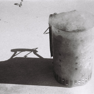 Op de afbeelding: Een zwarte metalen prullenbak met een deksel en gaten aan de zijkant. De prullenbak staat op een wit oppervlak.