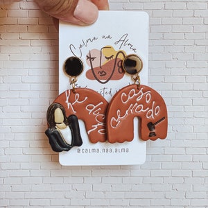 May include: A pair of terracotta-colored clay earrings with white lettering. The left earring features a woman's face and the text "He dicho". The right earring features a gavel and the text "caso cerrado". The earrings are on a white card with the text "Calma na Alma" and "@calma.naa.alma".