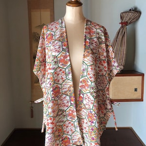 Peut inclure: Veste de style kimono à motifs présentée sur un mannequin. La veste présente un fond blanc avec un motif hexagonal répété rempli de motifs floraux et géométriques orange, rouges, violets et verts. La veste a des attaches à la taille.