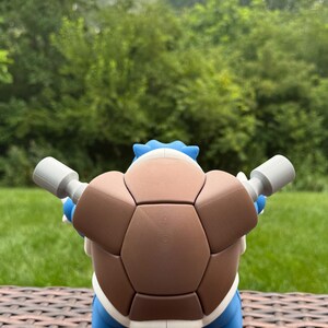 May include: A blue and brown turtle-like creature figurine featuring a large, segmented brown shell on its back, two grey cylindrical cannons extending from the sides, and a pointed blue tail visible at the bottom, viewed from the rear.