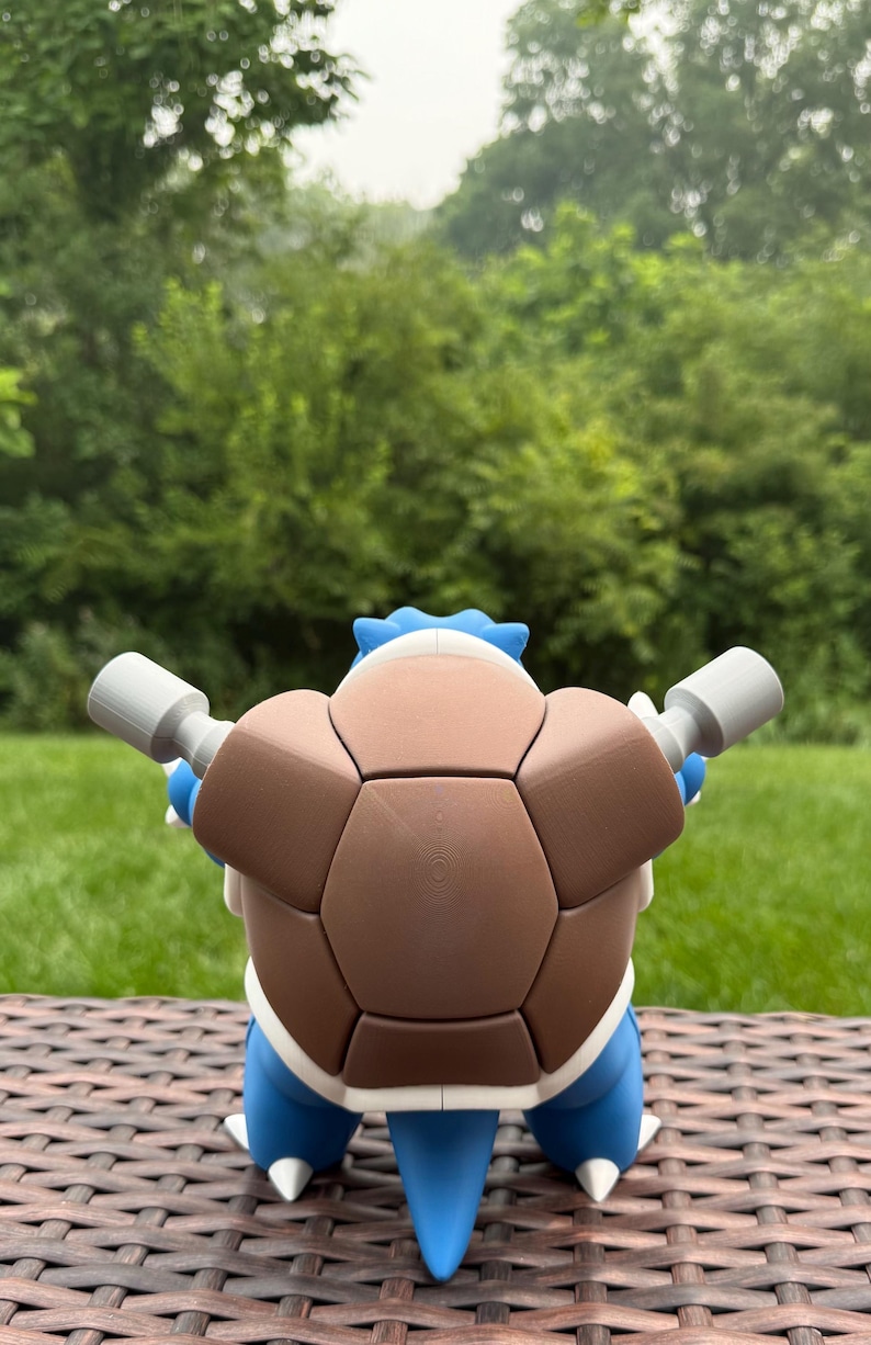 May include: A blue and brown turtle-like creature figurine featuring a large, segmented brown shell on its back, two grey cylindrical cannons extending from the sides, and a pointed blue tail visible at the bottom, viewed from the rear.
