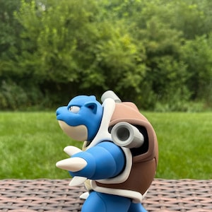 May include: A blue and brown bipedal creature figurine with a turtle-like shell, featuring two grey cylindrical cannons protruding from the back, a cream-colored underbelly, and sharp white claws on the hands and feet, captured in profile.