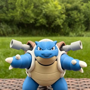 Puede incluir: Una figura de Blastoise azul y beige con dos cañones grises montados en su caparazón. El personaje tiene una postura bípeda, garras blancas en manos y pies, y un vientre color crema. La figura muestra capas de impresión 3D visibles, con una cabeza detallada de orejas puntiagudas y una expresión concentrada.