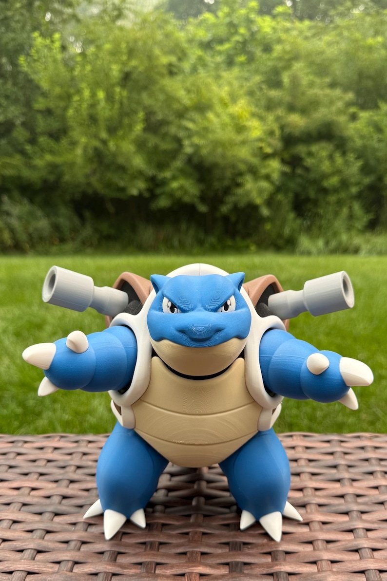 May include: A blue and beige Blastoise figurine featuring two gray cannons mounted on its shell. The character has a bipedal stance, white claws on its hands and feet, and a cream-colored underbelly. The figure is constructed with visible 3D printed layers, showcasing a detailed head with pointed ears and a focused expression.