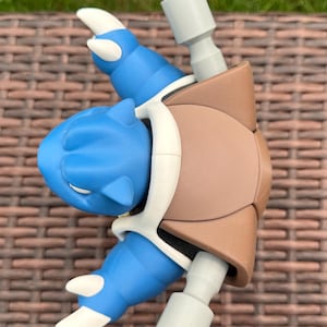 May include: A blue and brown plastic figurine featuring a creature with a large brown shell on its back, blue limbs with white claws, and two gray cylindrical cannons extending from the shell. The figure has a smooth matte finish.