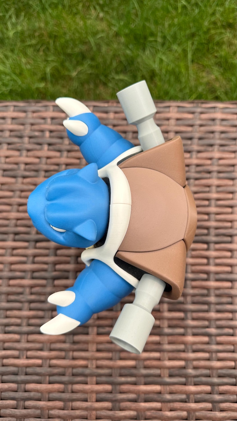 May include: A blue and brown plastic figurine featuring a creature with a large brown shell on its back, blue limbs with white claws, and two gray cylindrical cannons extending from the shell. The figure has a smooth matte finish.