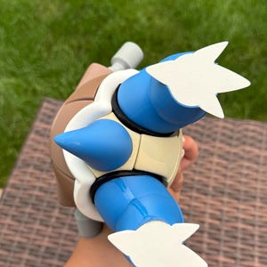 May include: A blue and tan plastic figure featuring a brown shell, a light-colored chest plate, and two blue limbs with white three-toed feet. The figure has a prominent blue spike on the chest and a gray cylindrical component extending from the back.