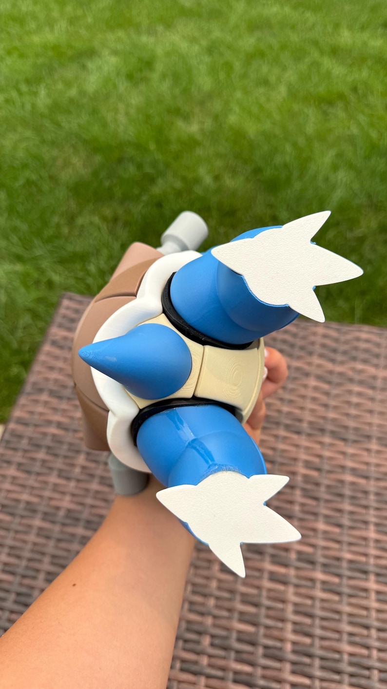 May include: A blue and tan plastic figure featuring a brown shell, a light-colored chest plate, and two blue limbs with white three-toed feet. The figure has a prominent blue spike on the chest and a gray cylindrical component extending from the back.