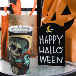 May include: A black and white stainless steel tumbler with a graphic of Jack Skellington and Sally from the Nightmare Before Christmas. The tumbler is sitting on a table with a sign that says "Happy Halloweeen".