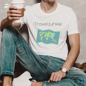 Puede incluir: Camiseta blanca con el texto "Travel Junkie" y una imagen de dibujos animados de un mapa del mundo doblado con alfileres rojos.