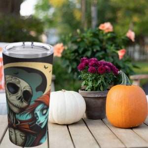 May include: A stainless steel tumbler with a black and white image of Jack Skellington and Sally from the Nightmare Before Christmas. The tumbler is sitting on a wooden table with a white pumpkin, an orange pumpkin, and a potted plant with purple flowers.
