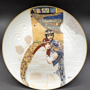 Puede incluir: Un plato de cerámica blanco con borde dorado presenta una pintura colorida de una pareja sosteniendo un bebé. La pintura está en un estilo que recuerda la obra de Gustav Klimt, con acentos de pan de oro y patrones geométricos.