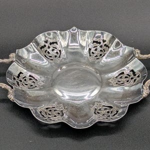 Puede incluir: Un plato plateado y ornamentado, con borde festoneado y dos asas. El plato presenta patrones florales decorativos recortados alrededor del borde. El centro del plato es liso y reflectante. Esta pieza decorativa es probablemente para servir o exhibir.