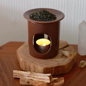 May include: A brown ceramic essential oil diffuser with a round top holding dried herbs. A lit candle is inside the diffuser, visible through a curved opening. The diffuser sits on a wooden slab, with pieces of wood and a crystal nearby.