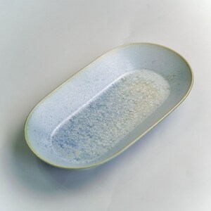 May include: A light blue oval serving platter with a speckled, textured surface. The ceramic dish has a slightly raised rim with a pale green edge. The interior has a gradient of blue and white tones.
