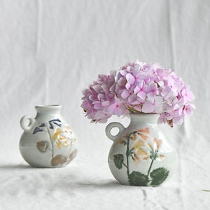 May include: Two small, off-white ceramic vases with floral designs and small handles. One vase holds a bouquet of pink hydrangeas. The vases are decorated with yellow, orange, and blue floral patterns and green leaves.