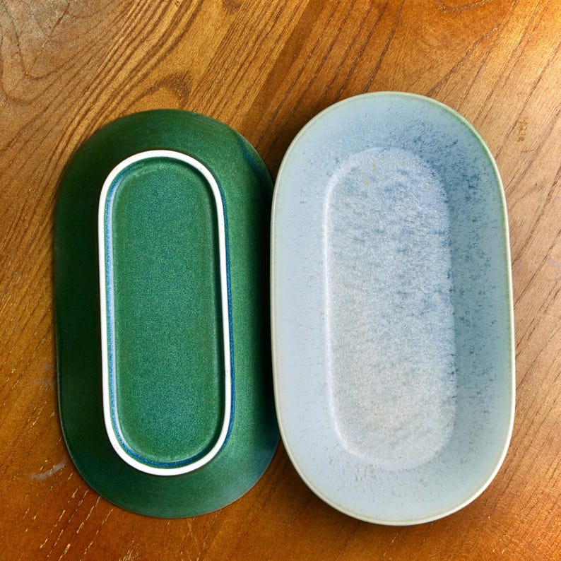 May include: Two oval-shaped ceramic serving platters. One is a deep green with a white rim, the other is a light blue with a speckled texture. Both are on a wooden surface.