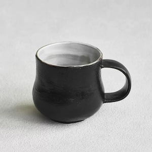K&ouml;nnte beinhalten: Eine wei&szlig;e Keramik-Tasse mit schwarzem Henkel und dem Text "But First, Coffee" in handschriftlichem Stil. Die Tasse steht auf einer wei&szlig;en Oberfl&auml;che. Die Tasse ist etwa 10 cm hoch.