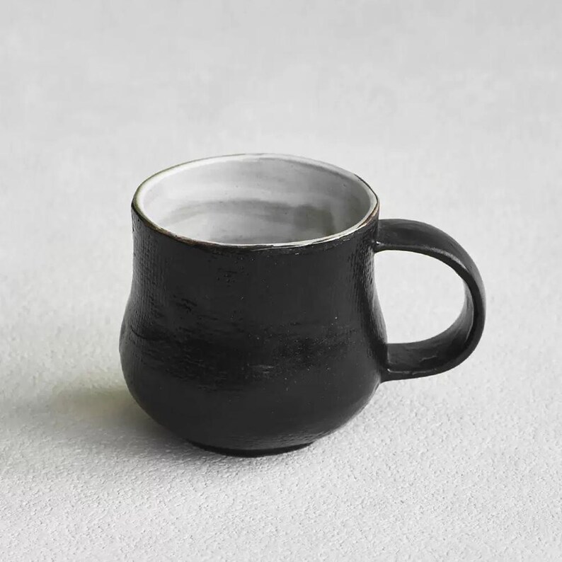 K&ouml;nnte beinhalten: Eine wei&szlig;e Keramik-Tasse mit schwarzem Henkel und dem Text "But First, Coffee" in handschriftlichem Stil. Die Tasse steht auf einer wei&szlig;en Oberfl&auml;che. Die Tasse ist etwa 10 cm hoch.