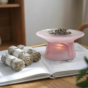 Op de afbeelding: Een roze etherische olie diffuser met gedroogde kruiden erop staat op een open boek. Bundels witte salie liggen naast de diffuser. De diffuser heeft een doorzichtige glazen bovenkant en een matroze basis.
