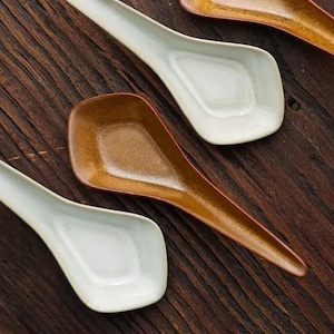 Puede incluir: Cuatro cucharas de cerámica, dos marrones y dos blancas, están dispuestas sobre una superficie de madera. Las cucharas son todas ligeramente curvadas y tienen un cuenco redondeado.